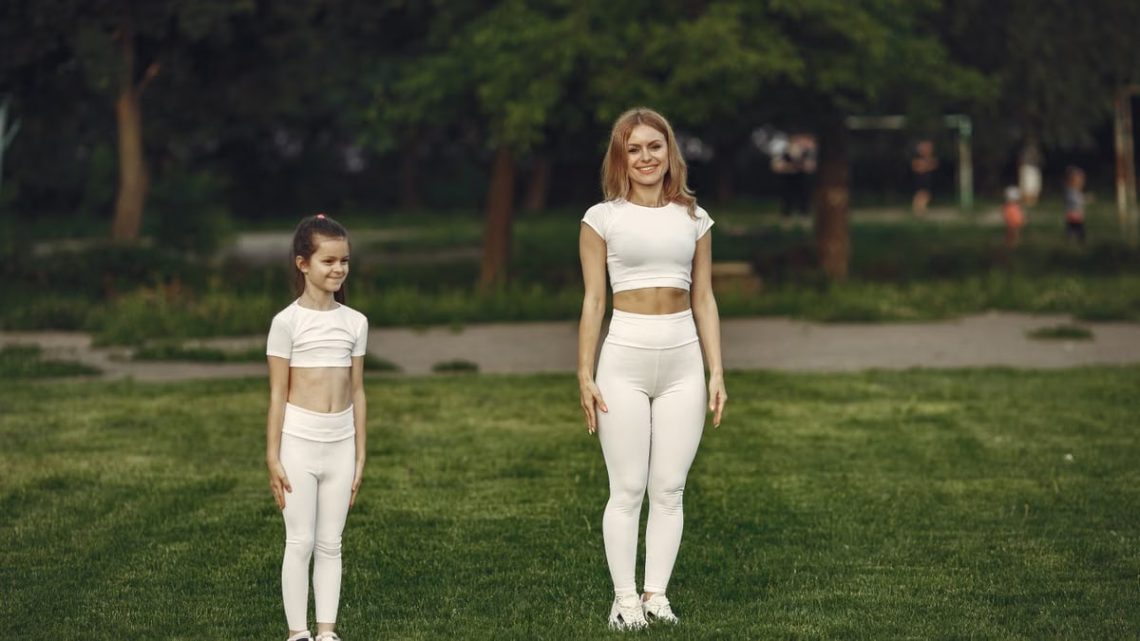 A woman and girl in white activewear standing on a grass field outdoors, smiling.